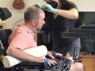 A man in a wheel chair is having a wire connected to hardware on his skull by a woman wearing medical gloves and a surgical mask.